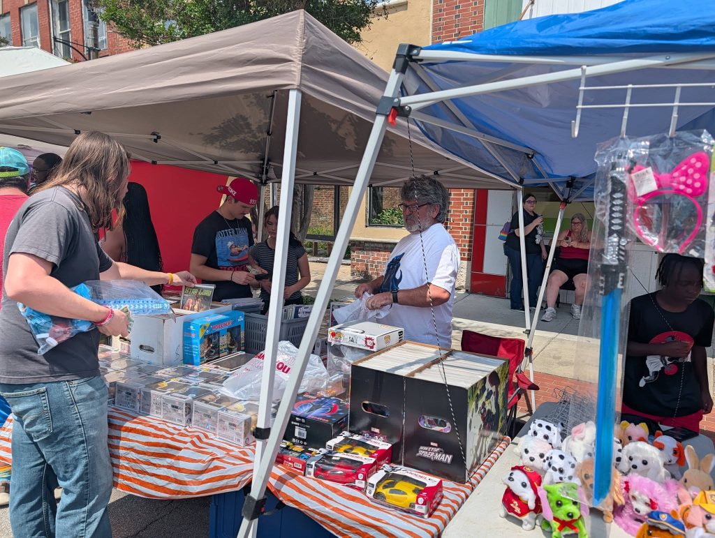 People browsing toys, collectibles, and stuffed animals at outdoor market booths on a sunny day, with several people sitting in the background.