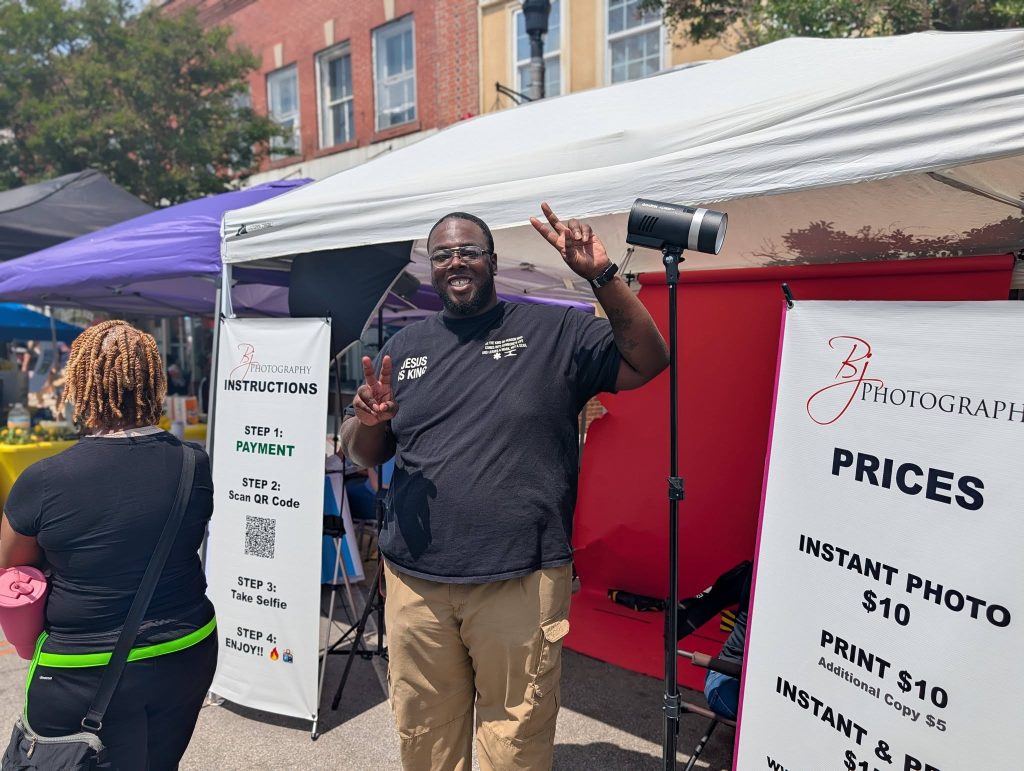 Man smiling and flashing peace signs at photography booth outdoors, surrounded by banners displaying instructions and prices for instant photos; another person stands nearby.