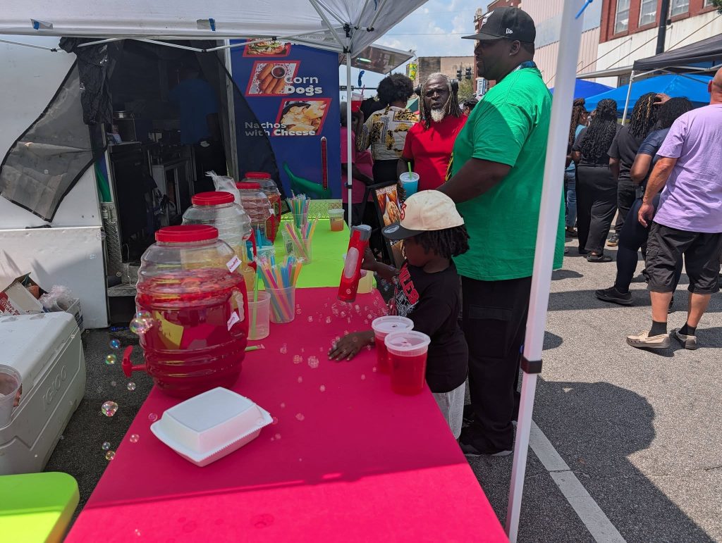 People standing at a vendor booth with drink dispensers, colorful straws, and food containers at an outdoor street festival on a sunny day.