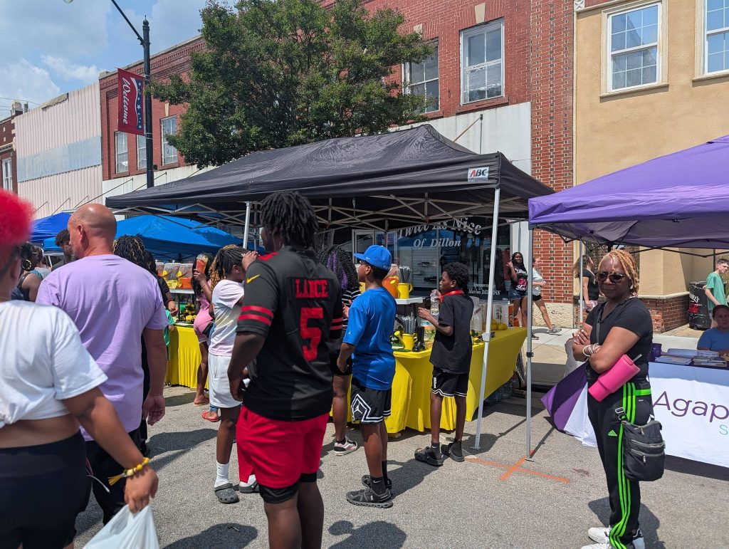 People gather at outdoor market stalls on a sunny day, with tents and tables set up along a city street lined with brick buildings.