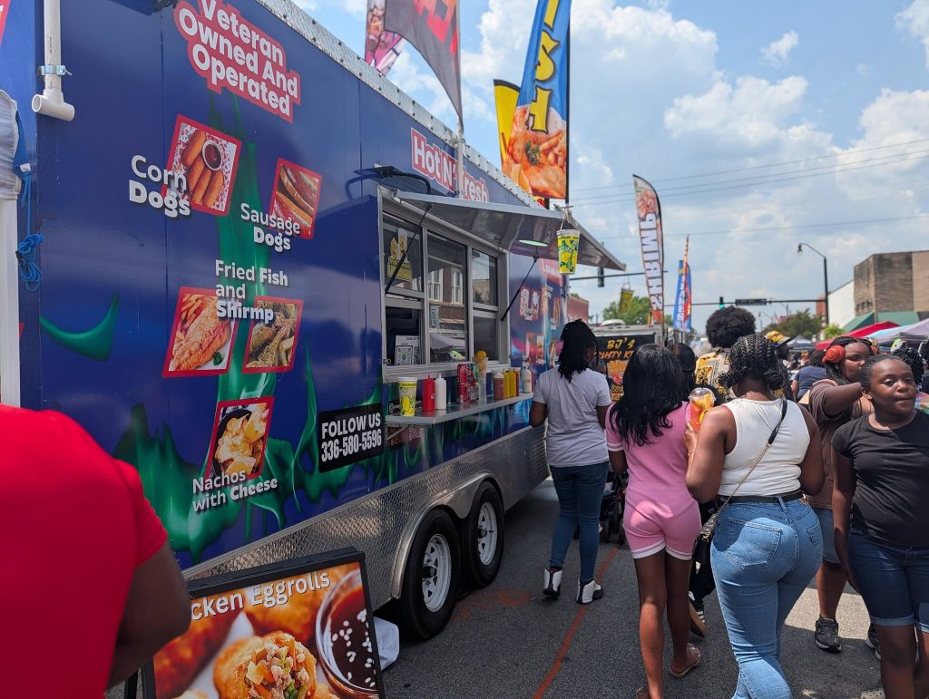 People standing in line at a food truck offering corn dogs, sausage dogs, fried fish, shrimp, nachos with cheese, and chicken eggrolls at an outdoor event.