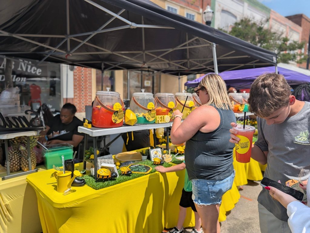 People buying drinks at a vibrant outdoor lemonade stand with yellow tablecloths, colorful drink dispensers, and cheerful decorations on a sunny day.