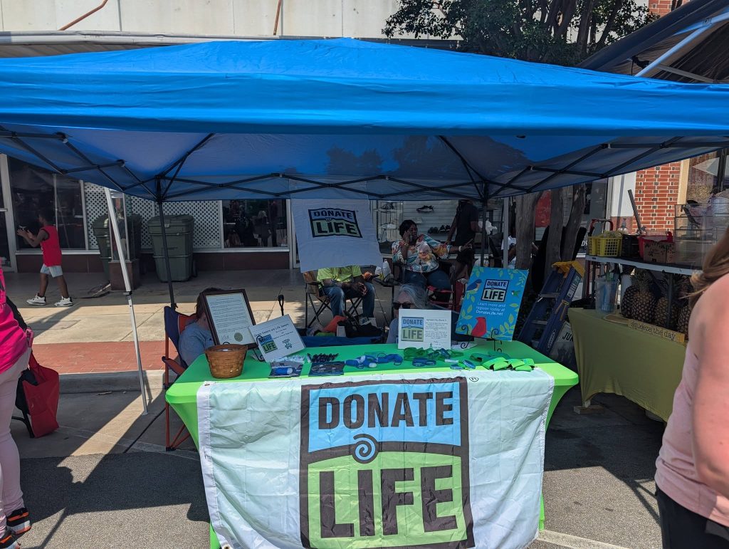 A "Donate Life" booth at an outdoor event with informational materials, a tablecloth banner, and people sitting under a blue canopy.