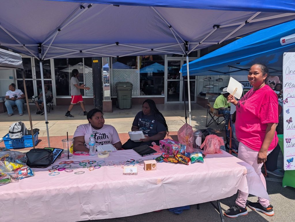 Three women at an outdoor booth selling bracelets, snacks, and drinks, under a blue canopy at a street fair. One woman stands holding a fan.