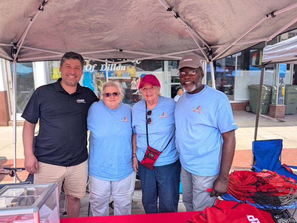 Four adults, three wearing blue shirts, stand and smile under a canopy at an outdoor event, with AARP branding and giveaway items visible on the table.