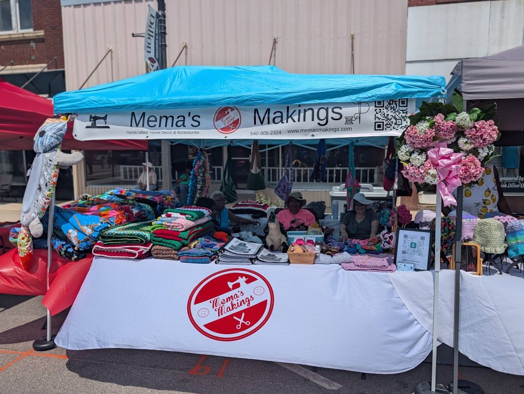 Outdoor market stall "Mema's Makings" with colorful handmade blankets, crocheted items, plush toys, and decorative wreaths, run by two women under a blue canopy.