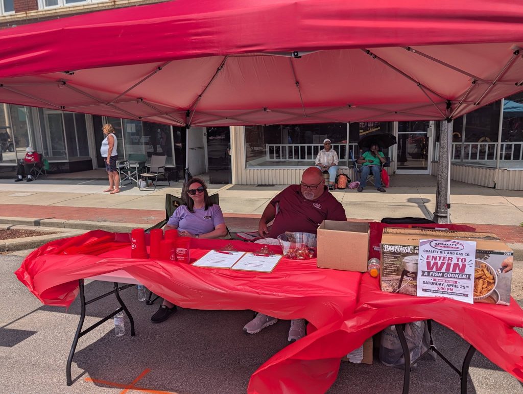 Two people sit under a red canopy at a street booth covered with red cloth, promotional cups, raffle sign, and entry forms, with pedestrians and seated people in the background.
