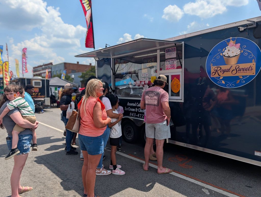 People wait in line at the Royal Sweets food truck on a sunny day at an outdoor festival.