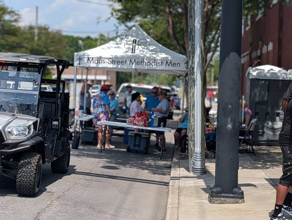 A group of people gather under a tent labeled "Main Street Methodist Men" at an outdoor event, with a utility vehicle parked nearby and a person walking on the sidewalk.