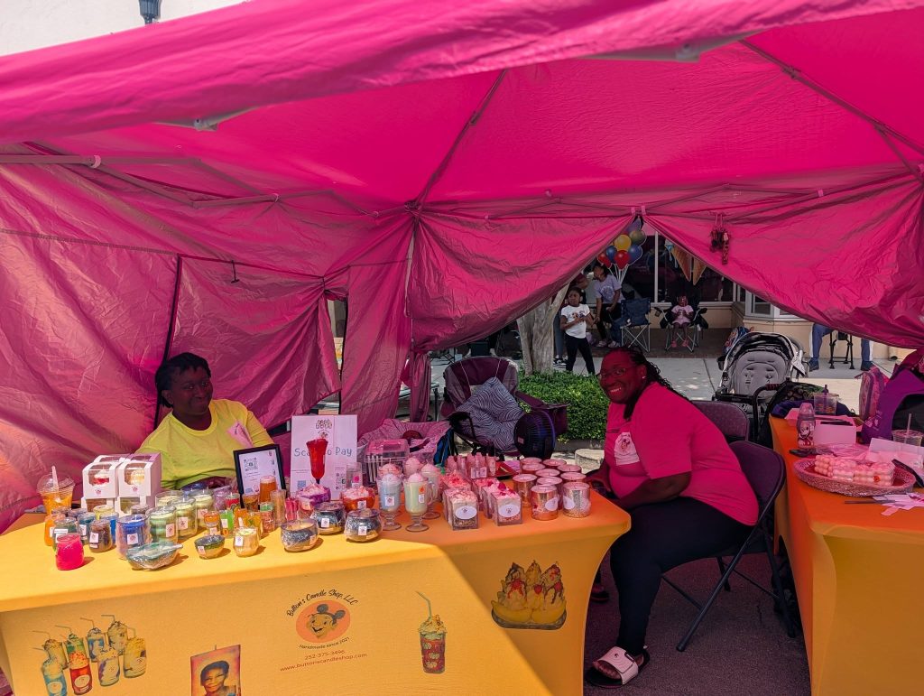 Two women sit under a pink canopy at a market stall displaying candles and other products on yellow tablecloths, with people and balloons visible in the background.