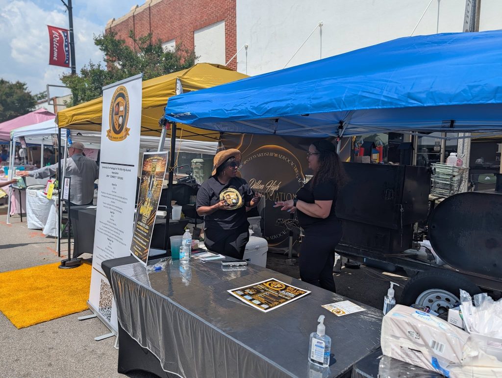Two women converse under a blue canopy at an outdoor event, surrounded by promotional materials, a grill, and hand sanitizers on a table.