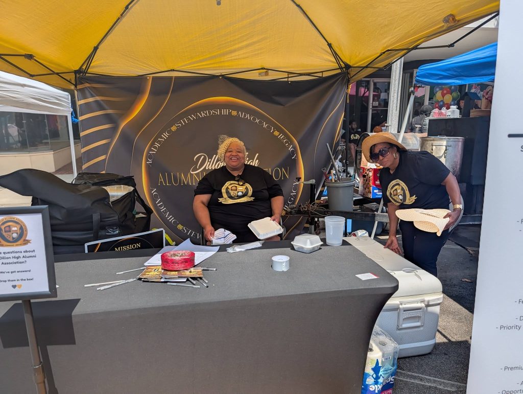 Two women at a booth under a yellow tent, smiling, with Dillon High Alumni Association sign and items on the table.