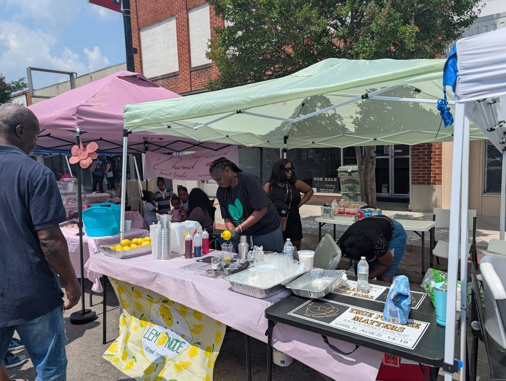 Outdoor street market with people selling lemonade and snacks under tents, tables covered with supplies, and a lemonade banner displayed in front.