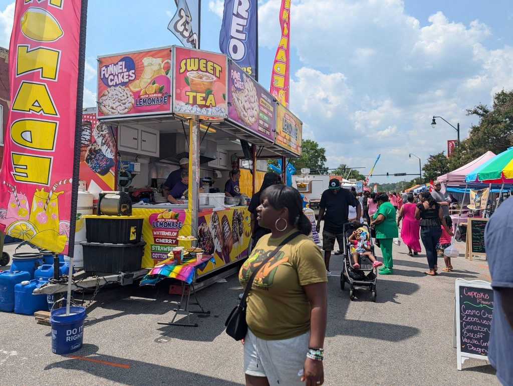 People walk past colorful food stalls at an outdoor street fair, with signs advertising lemonade, funnel cakes, and sweet tea on a sunny day.
