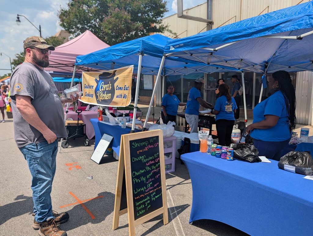 Man orders food at an outdoor street market booth with a blue tent, "Cora's Kitchen" sign, menu board, and several people in matching blue shirts serving food.