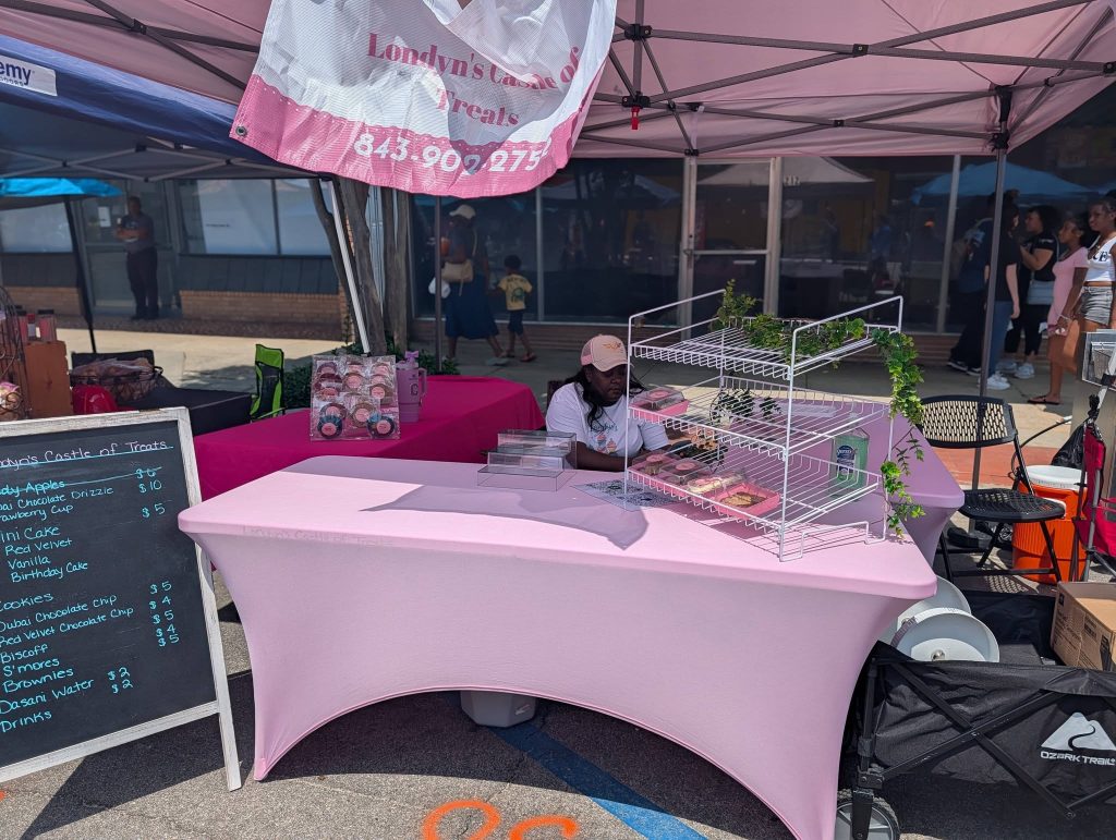 A vendor sits at a pink-covered booth selling treats at an outdoor market, with a menu board and display shelves of desserts under a canopy.