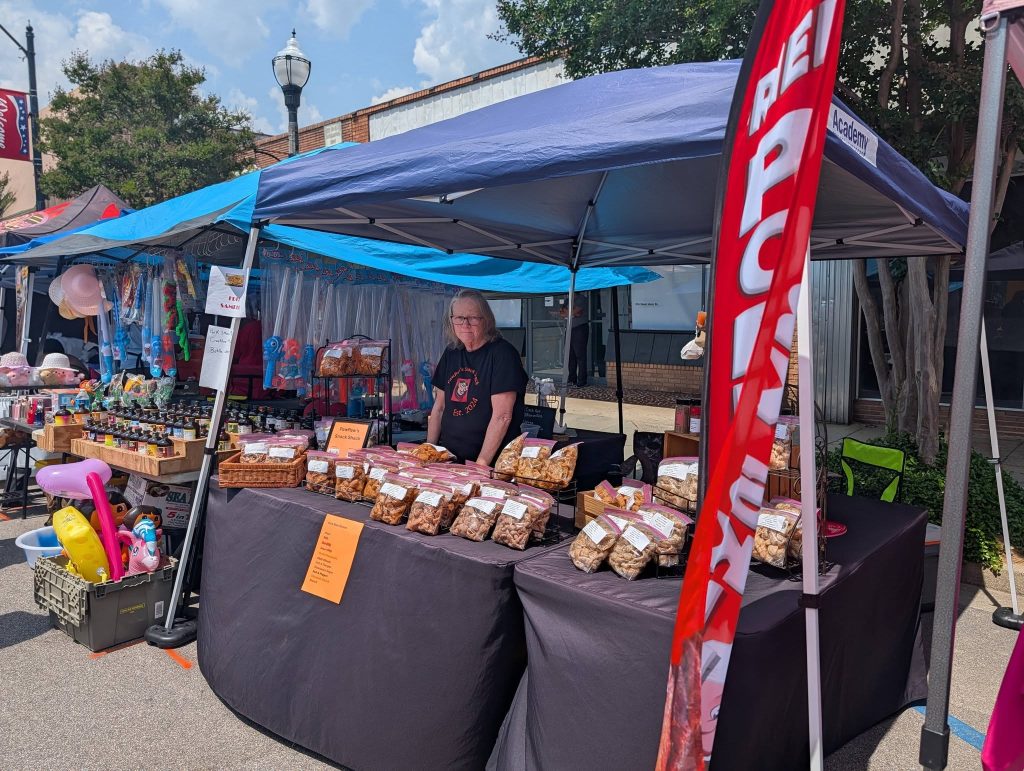 A person stands behind a market stall under a blue canopy, selling various packaged snacks and baked goods at an outdoor street fair.