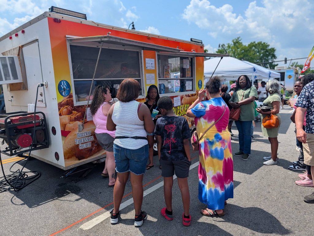People standing in line at a food truck on a sunny day, with tents and more people in the background at an outdoor event.