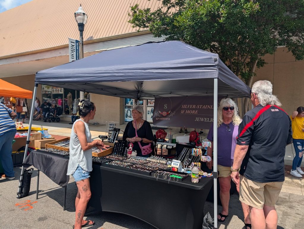 People shopping at an outdoor market jewelry stall under a canopy, with various jewelry items displayed on tables and others browsing in the background.