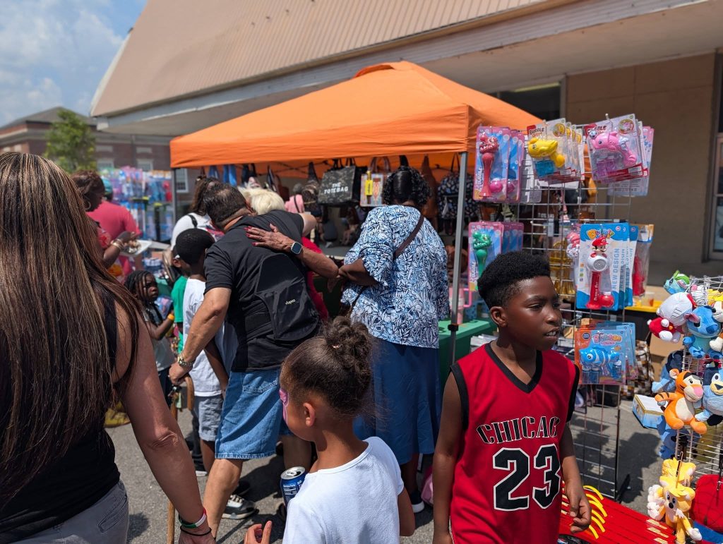 People gather at an outdoor market booth with toys and stuffed animals displayed, under an orange canopy on a sunny day.
