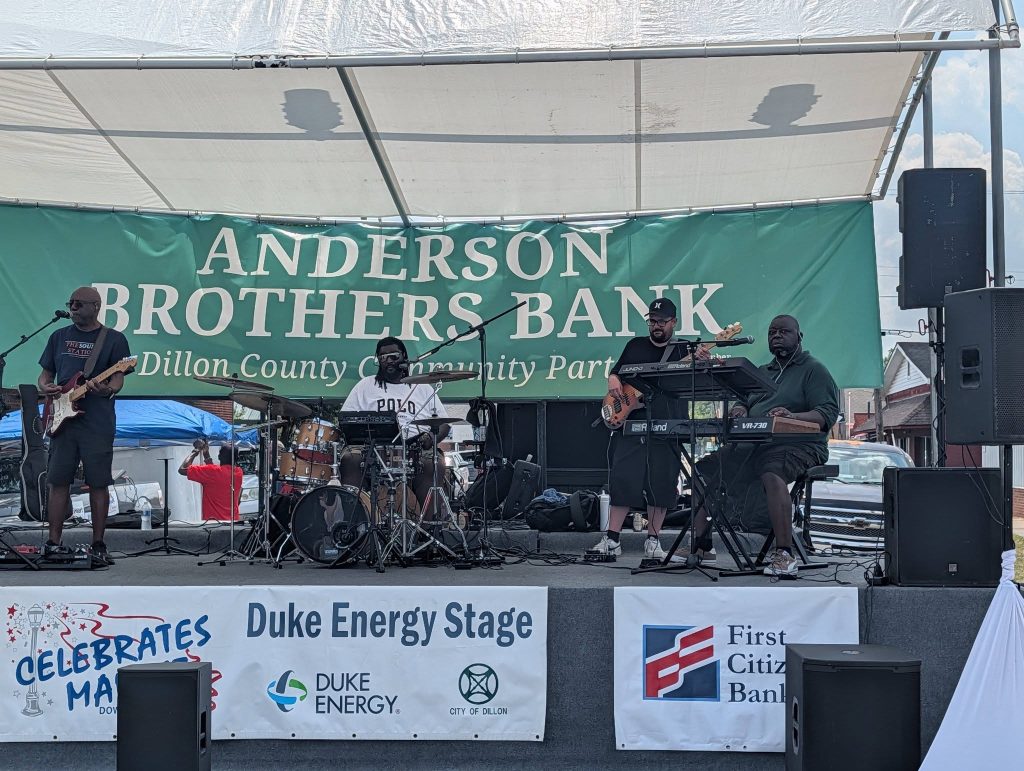 Four musicians perform on an outdoor stage with instruments under a tent, with banners and a crowd visible in the background.