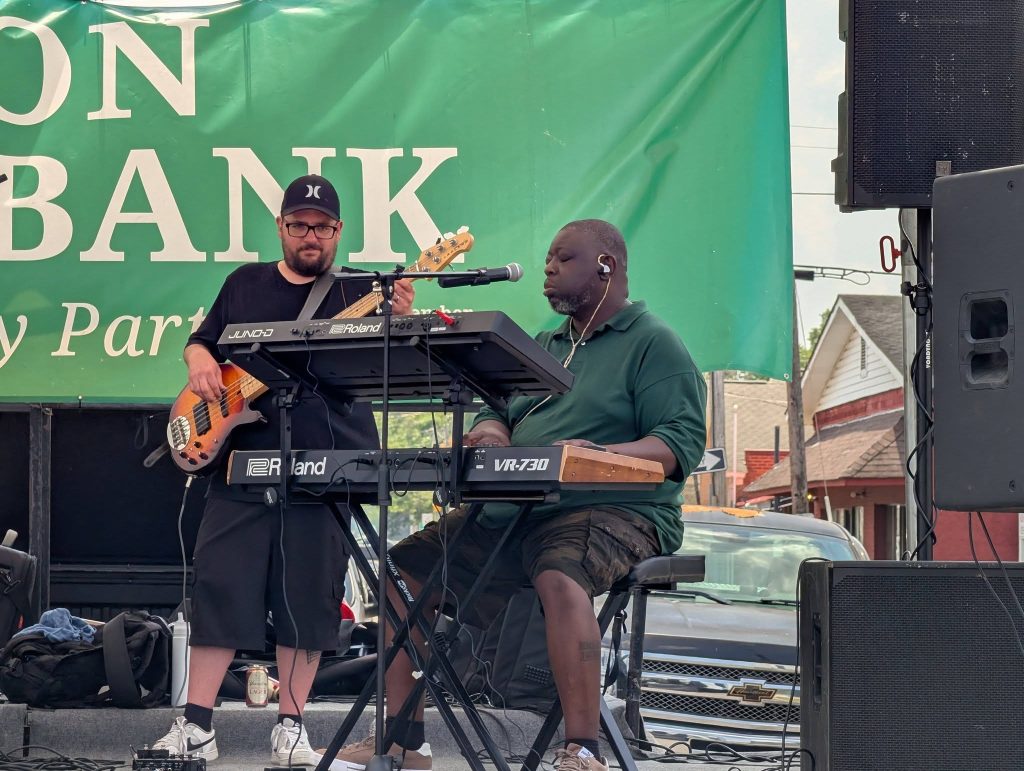 Two musicians performing outdoors, one playing bass guitar and the other on keyboard, with a green banner and houses in the background.