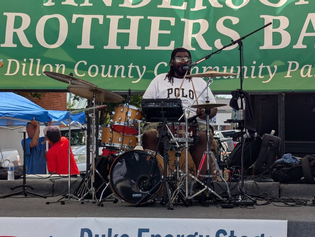 A drummer performs on stage, singing into a microphone, with drum kit and equipment, at an outdoor event under a green banner.