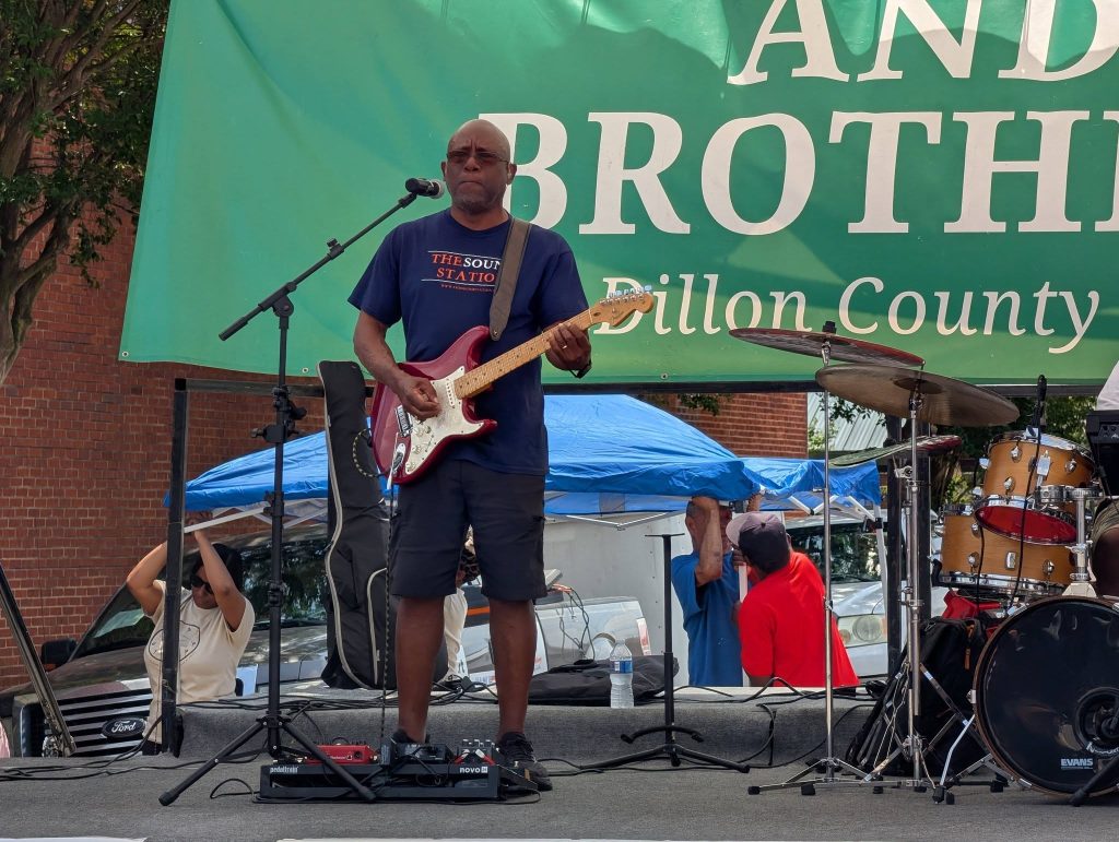 A man playing electric guitar on an outdoor stage, next to a drum set, with people and tents in the background during a live event.