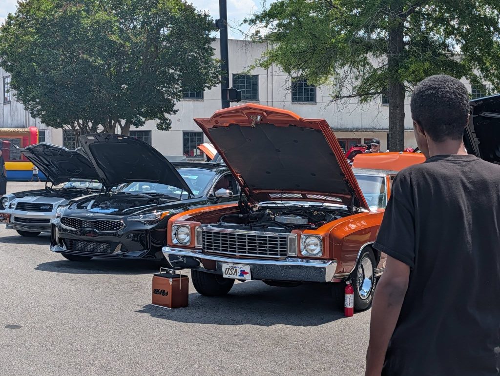 Several cars with open hoods displayed at a car show, people observing, trees and a building in the background, and a person in the foreground.