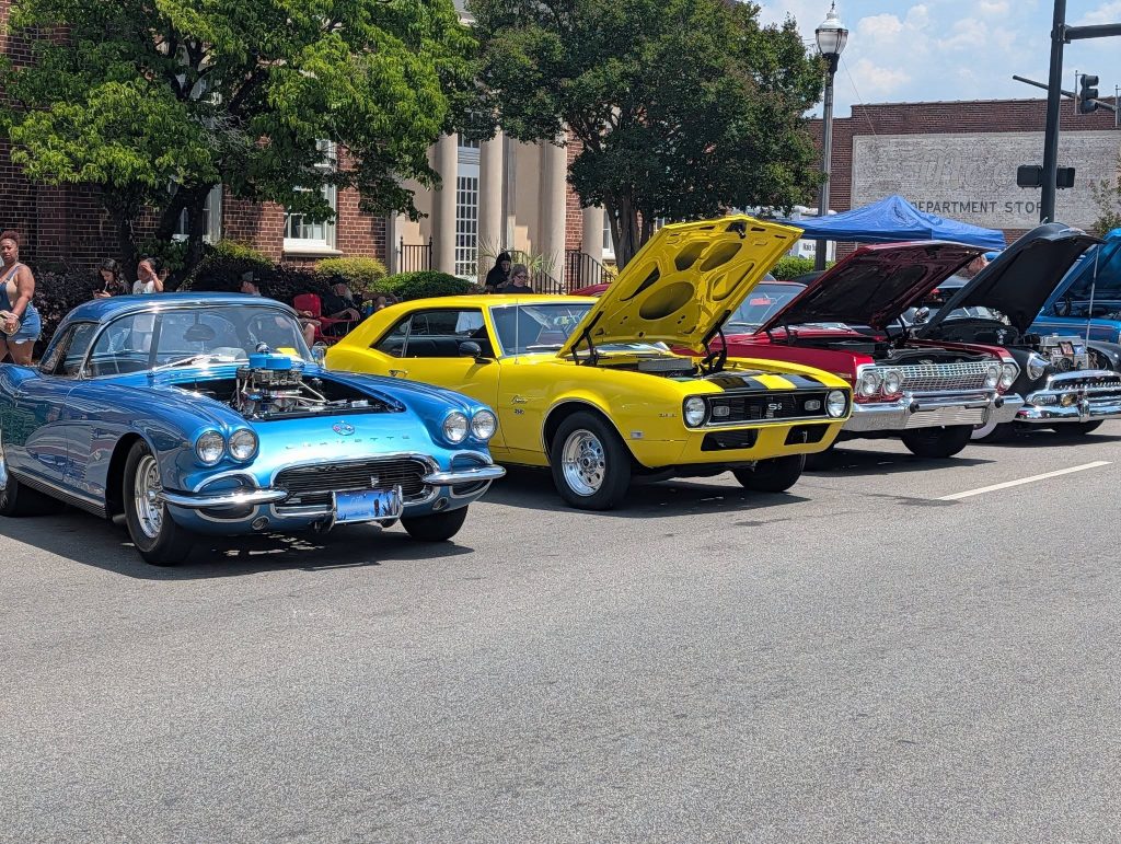 Classic cars with open hoods on display at an outdoor car show, with people in the background and trees lining the street.