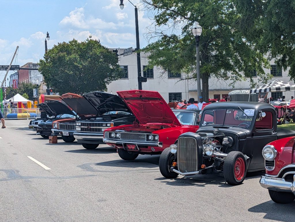 Classic cars with open hoods lined up at an outdoor car show, people in the background, with trees, tents, and buildings visible on a sunny day.