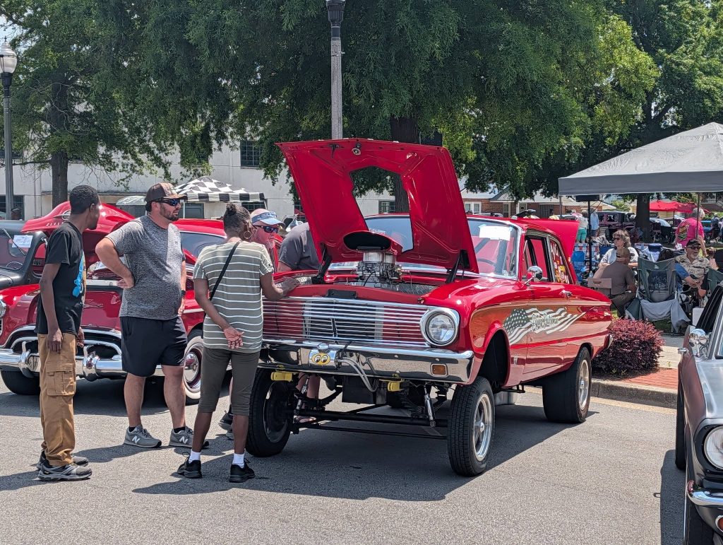 People gather around a red classic car with its hood open at an outdoor car show, surrounded by other vintage vehicles and trees.