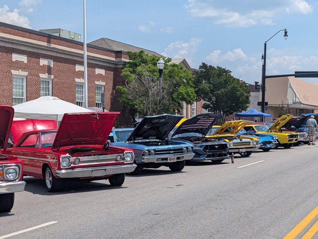 Classic cars with hoods open are lined up on a street for a car show, with people and tents visible in the background.