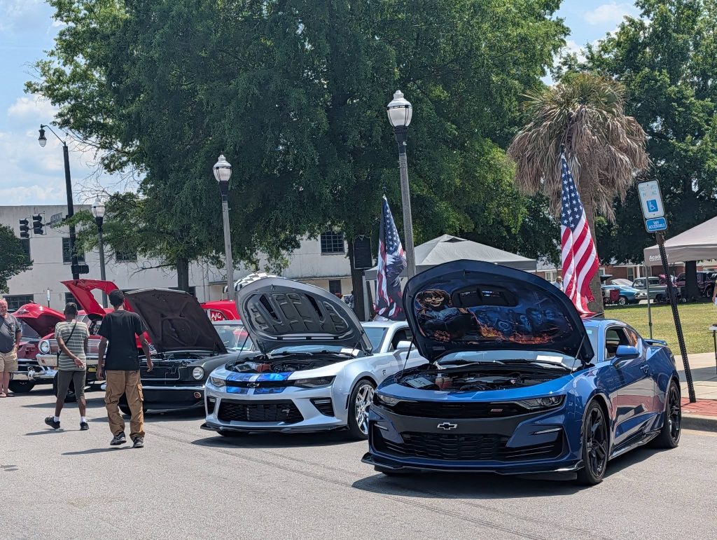 Several Chevrolet Camaros with open hoods on display at an outdoor car show, with American flags and people walking by on a sunny day.