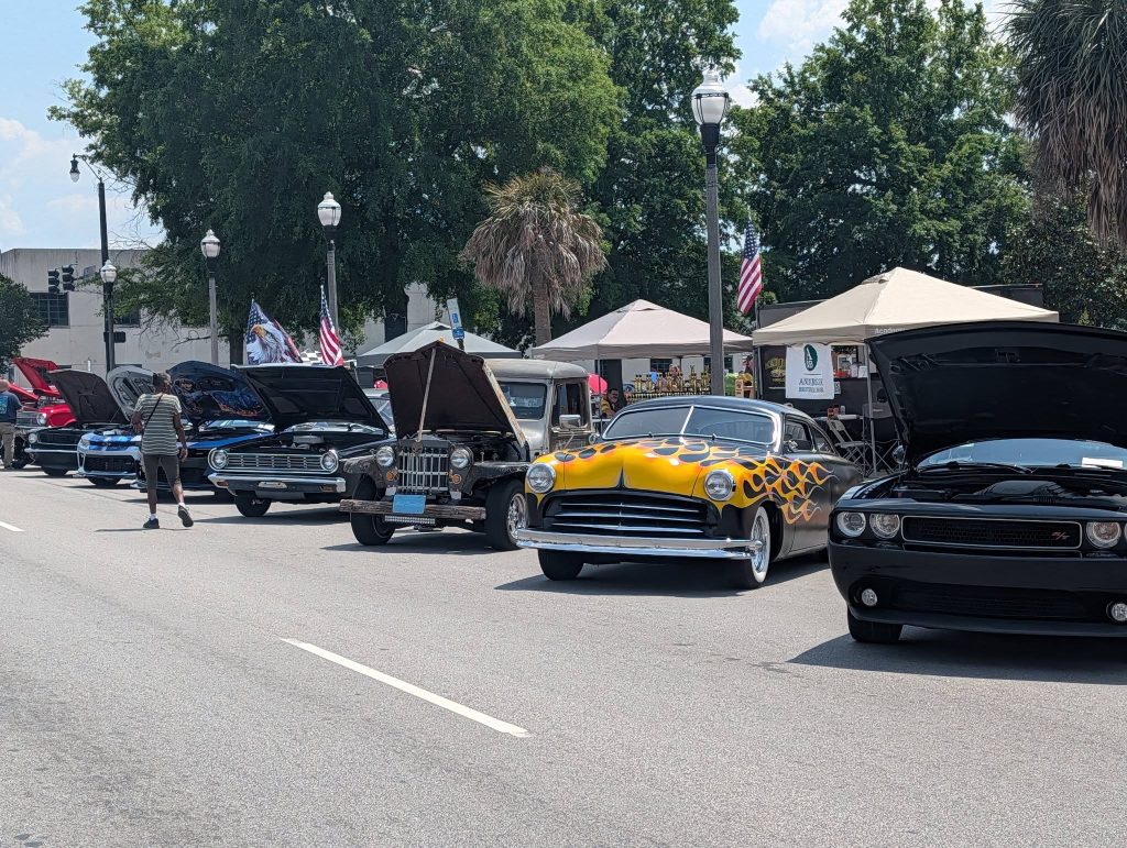Classic and modern cars with open hoods are displayed at a car show on a sunny street, with people walking and tents in the background.