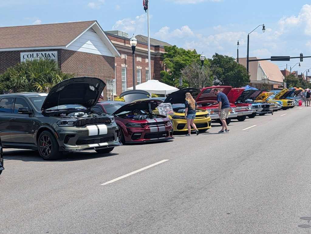 A row of colorful cars with open hoods displayed at a car show on a sunny street, people walking and observing vehicles.