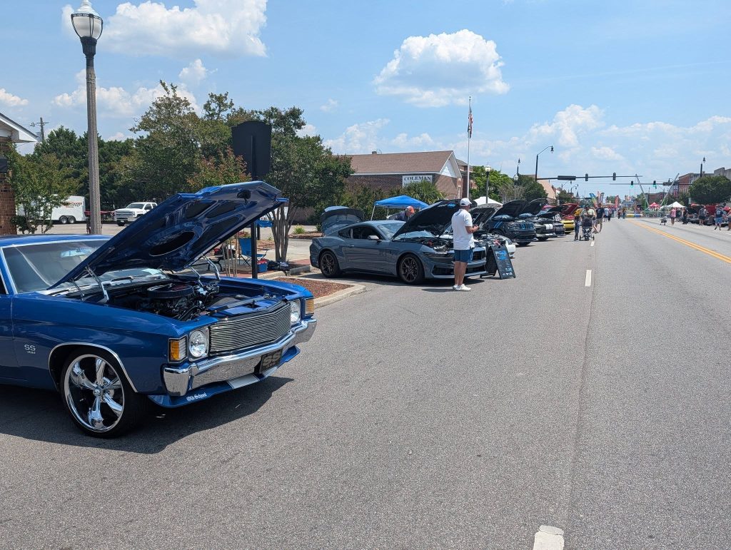 Classic and modern cars with open hoods lined up on a sunny street for a car show. People are observing and walking along the display.