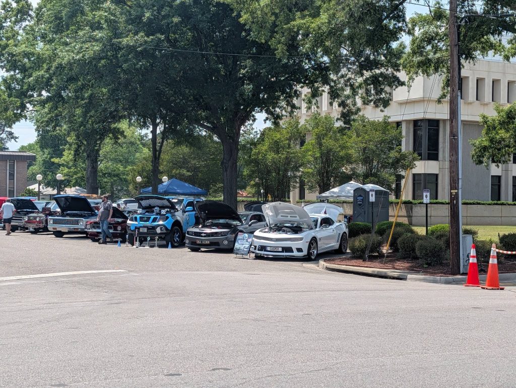 Several cars with open hoods are lined up at an outdoor car show, with people and tents under large trees near a modern building.
