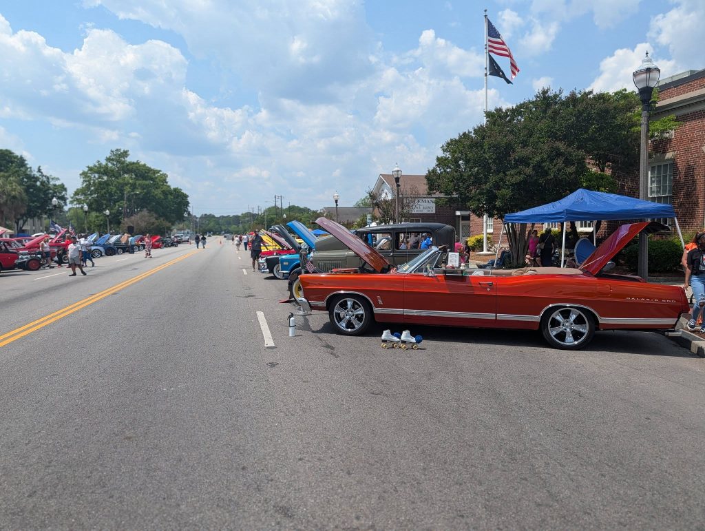 Classic cars with open hoods line both sides of a sunny street during a car show, people walking and viewing vehicles, American flag in the background.