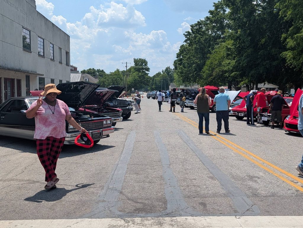 People walking and admiring classic cars displayed on both sides of a street during a sunny day, with one person posing and making a peace sign in the foreground.
