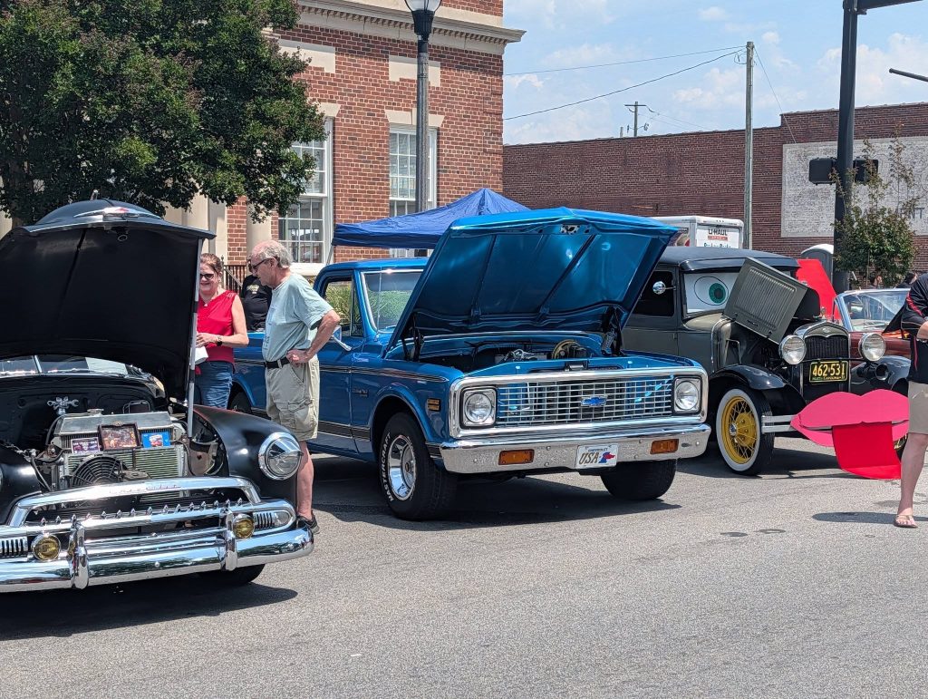 Classic cars with open hoods on display at an outdoor car show, people admiring the vehicles, brick buildings and blue sky in the background.