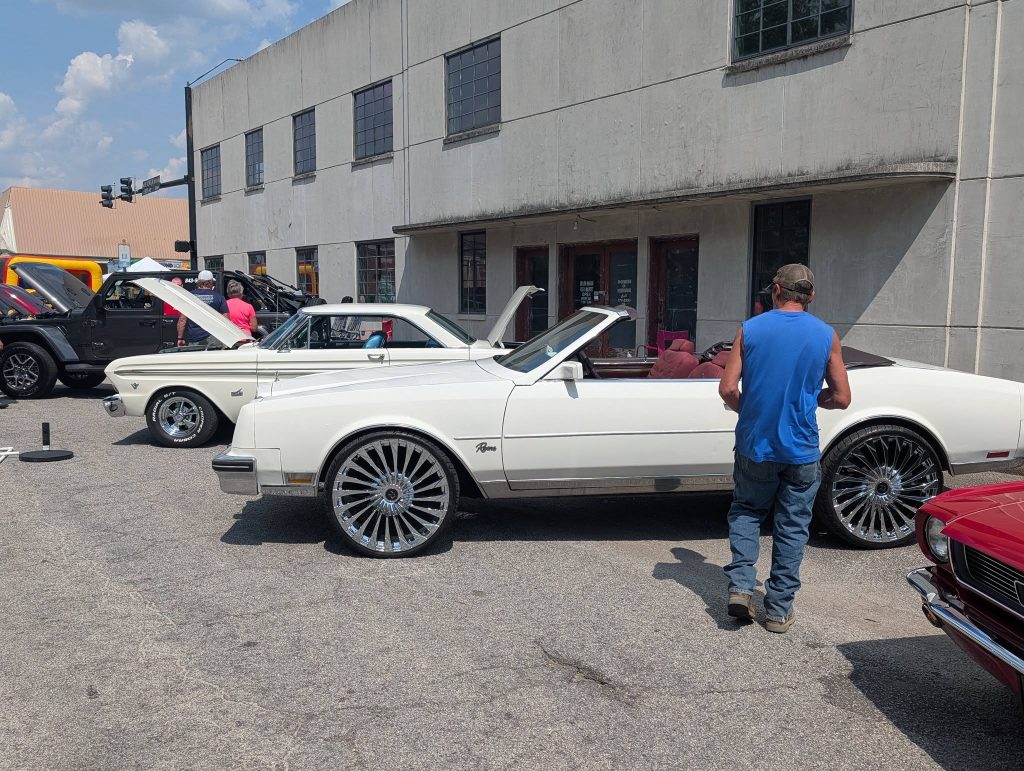 Several classic cars are parked on the street during a car show, with people walking and observing, in front of a building with large windows.
