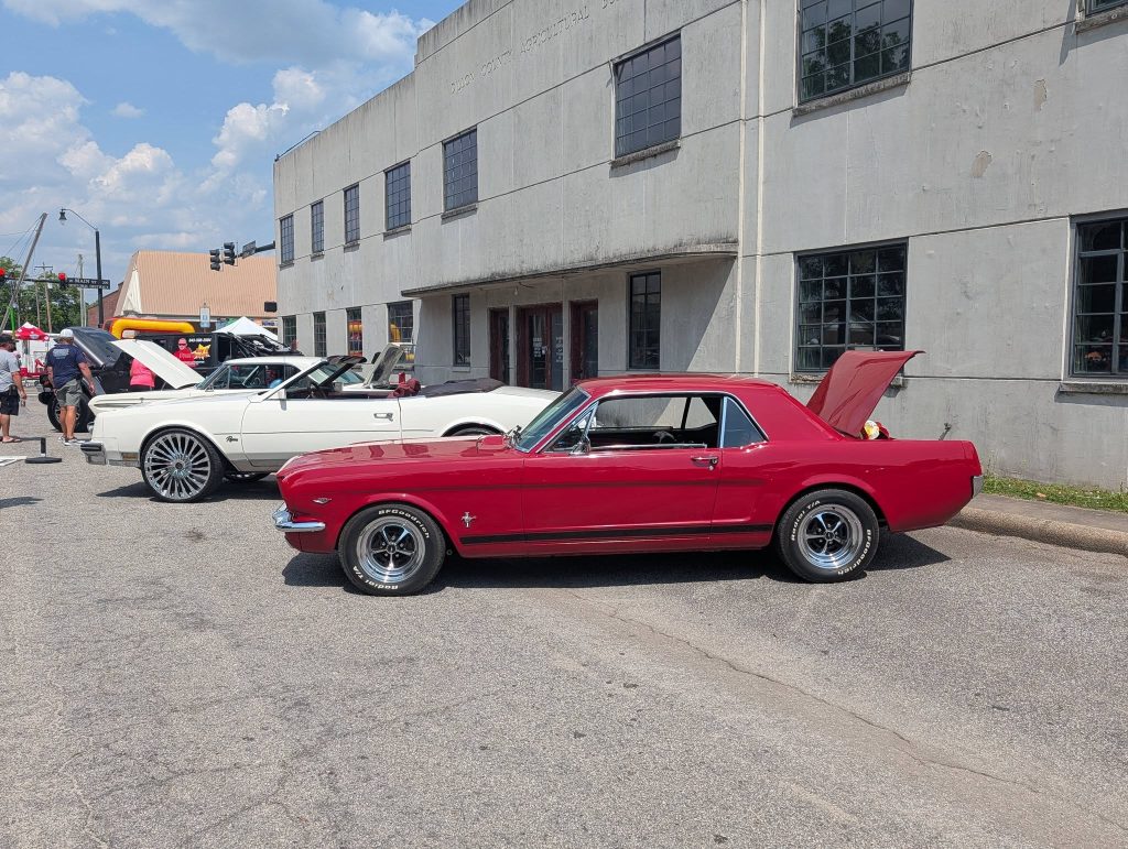 A red classic car with its trunk open parked beside a white convertible at an outdoor car show near a concrete building.