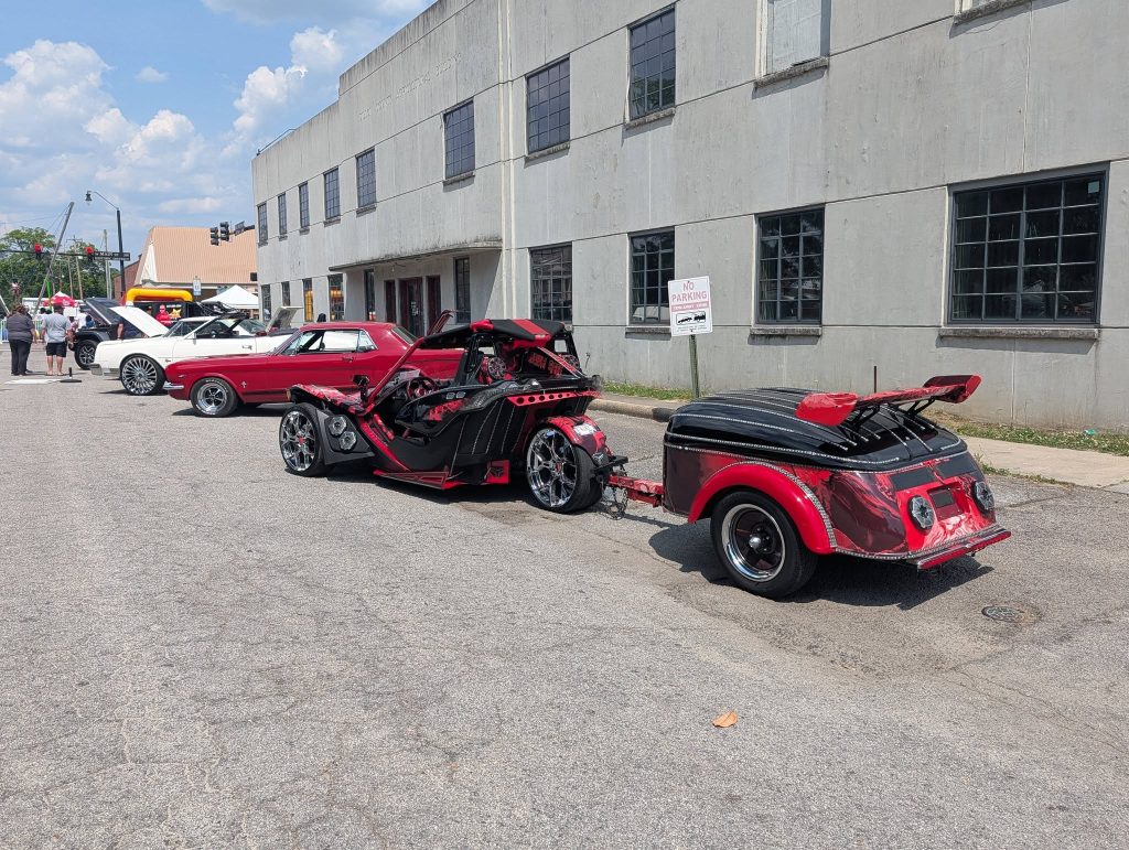 A row of classic and custom cars parked on a city street during a car show, with people walking nearby and a building in the background.