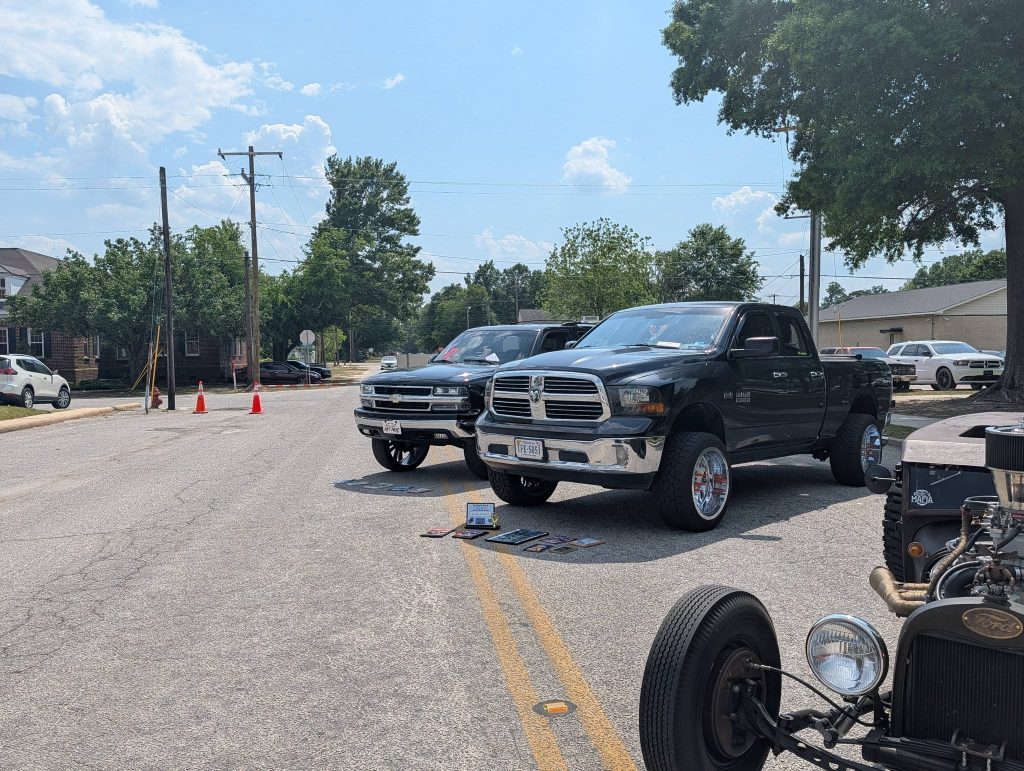 Two black pickup trucks and a vintage car parked on a sunny street with some tools and items displayed on the ground.