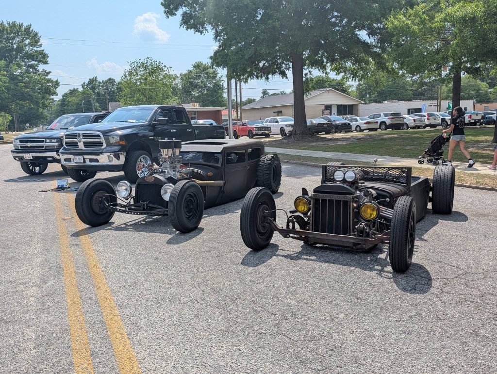 Two vintage rat rod cars parked on a street, with two modern black pickup trucks behind them and people walking in the background.