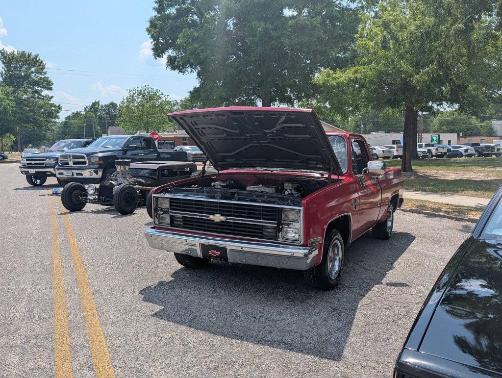 A red vintage Chevrolet pickup truck with its hood open, parked on a street among other classic vehicles at a car show under sunny weather.