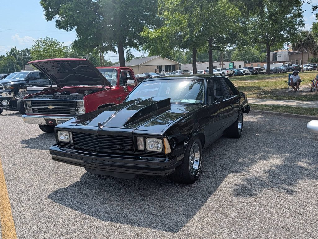 A black classic car with a raised hood scoop parked at a car show, with a red truck and other vehicles in the background.