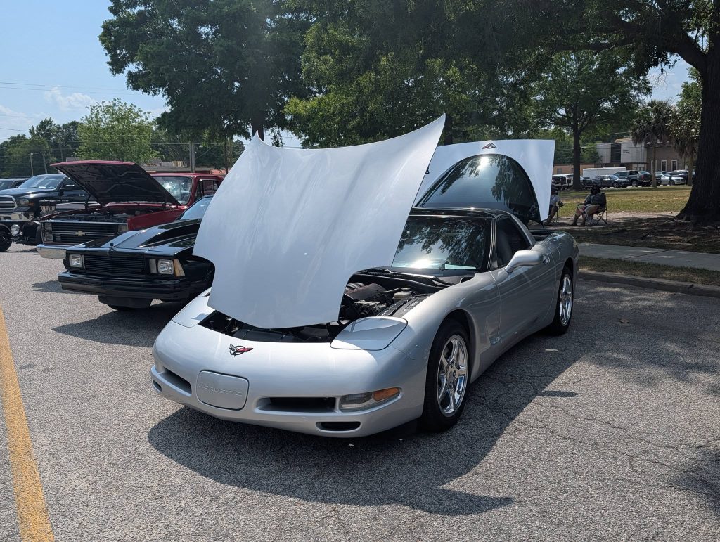 A silver Chevrolet Corvette with its hood open is parked at a car show, alongside other vintage vehicles, on a sunny day.