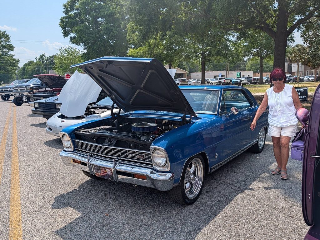 A row of classic cars with open hoods at a car show, a woman in white clothing stands next to a blue vintage car.